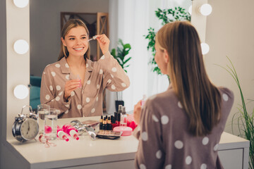 Young woman in polka dot pajamas applying cosmetics at a vanity mirror, smiling in natural daylight