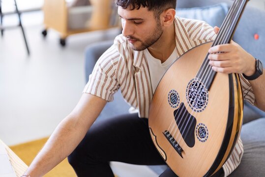 A young man with an oud sits at a table, focusing on writing music in a notebook, with a laptop and headphones nearby