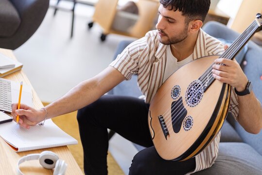 A young man with an oud sits at a table, focusing on writing music in a notebook, with a laptop and headphones nearby
