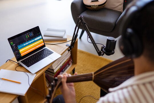 A musician is captured recording music in a home studio setup, with a microphone and a laptop displaying sound editing software