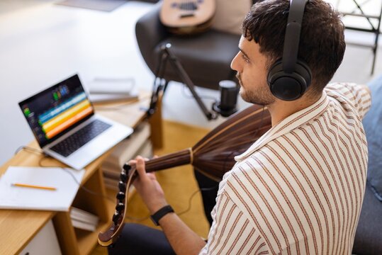 A musician is captured recording oud music in a home studio setup, with a microphone and a laptop displaying sound editing software