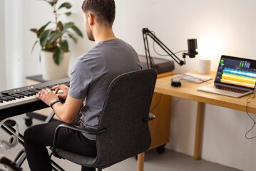 A man sits focused at a keyboard in a minimalist home studio setup, with a laptop showing audio tracks on the desk nearby, creating a dedicated space for sound editing and music production