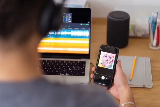 A man, seen from behind, holds a smartphone displaying colorful music graphics while using audio editing software on a laptop, seated in a modern workspace with additional audio devices
