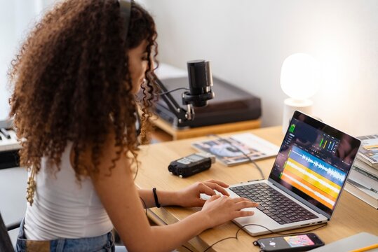 A young woman with curly hair and headphones focuses on recording music on her laptop, using a professional microphone and surrounded by audio equipment in a cozy, well-lit room