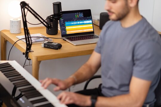 A man plays an electronic keyboard in a home recording studio, with a laptop displaying audio software and a microphone positioned on a nearby desk