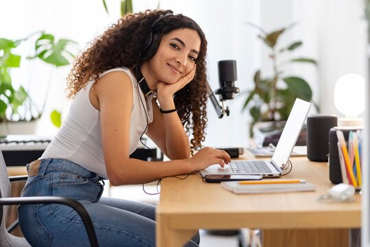A woman with curly hair wearing headphones focuses intently on a laptop while working at a desk setup with a microphone and various office supplies, surrounded by greenery - Powered by Adobe