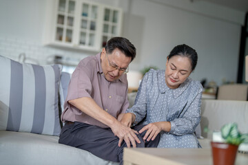 An elderly couple sits in their living room. The man holds his knee, appearing to be in discomfort, while his partner offers support. The image conveys care, concern, and the bond between the couple.