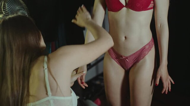 Hot italian professional female model in red panties and bra, while being smeared with skin oil by a fellow brunette model in a dressing room.