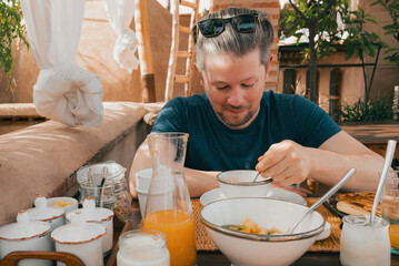 Young adult caucasian man eating a traditional Moroccan breakfast meal with fresh fruit juice, yogurt, bread in outdoor setting, local cuisine at a luxury riad hotel, Marrakech, Morocco, North Africa