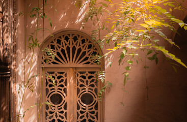 Ornate moroccan traditional wooden door with intricate lattice design, textured wall and surrounding greenery in a peaceful luxury riad courtyard with dappled sunlight, Medina, Marrakech, Morocco