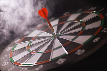 Fototapeta premium Red Dart Hitting Bullseye on Traditional Dartboard with Smoky Background