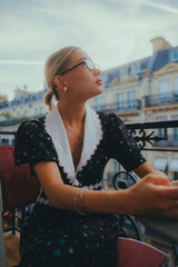Obraz premium Elegant woman with glasses sitting outdoors, wearing a vintage floral dress with lace details, against a backdrop of classic European architecture, showcasing beauty and sophistication.
