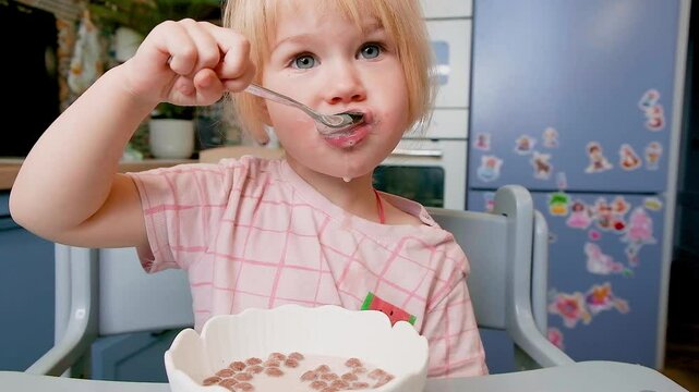 A little Caucasian child girl of two years old eats chocolate balls with milk for breakfast in the kitchen.