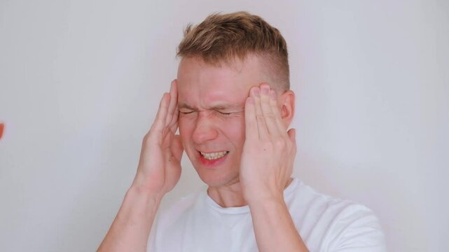 A young Caucasian man touches his head and winces in pain, the concept of migraines and headaches.