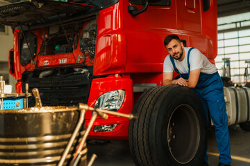 Fototapeta premium Mechanic in blue working suit leans on a large truck tire in a garage, with a red truck in the background.