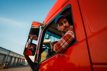 A truck driver leans out of the window of a red truck while parking on a parking lot in an...