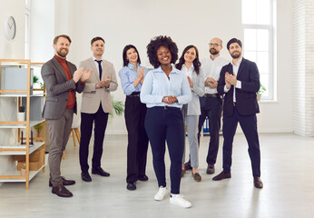 Happy company employees congratulating happy female colleague on job well done applauding standing in office. Business people and group of staff clapping to young african american woman.