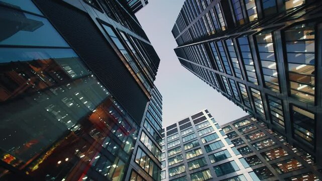 Wide angle gimbal dolly shot downtown high-rise glass skyscrapers in evening, financial district looking up tall modern architecture office buildings city center street, warm lighted windows