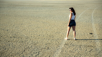 Young woman in shorts and sunglasses stands on dry desert ground.
