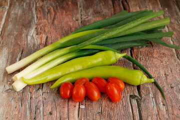 A fresh assortment of green onions, long green peppers, and ripe cherry tomatoes arranged on a rustic wooden surface. 