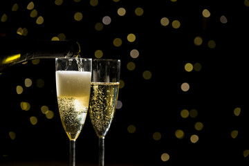 Pouring champagne into two glasses on a black background with golden bokeh lights, close up shot. New Year's Eve, happiness, and hope concepts.