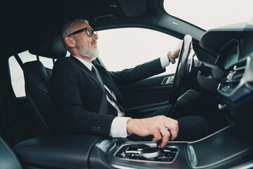 Mature businessman in formal suit driving car confidently on city street, highlighting professional...