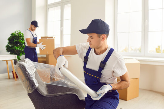 Young confident man mover packing furniture in the living room. Moving service men workers working in clients house wrapping chair with plastic wrap. Move, relocation and moving day concept.