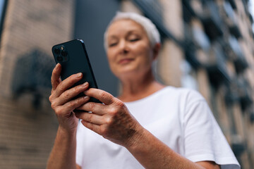 Selective focus of gray-haired senior lady using smartphone texting message standing on urban...