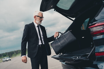 Senior businessman in formal suit retrieving bag from car trunk in urban parking setting
