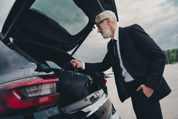 Senior businessman in formal wear loading suitcase into car on city street parking lot