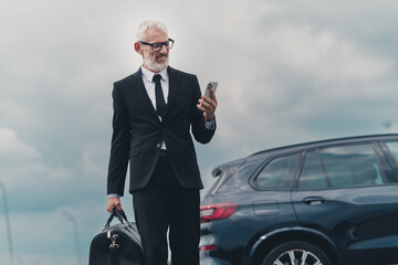 Senior businessman in formal suit outside on city street near car with smartphone and travel bag