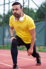 Active sports enthusiast in wireless headphones prepares leg muscles for training on rubberized running track at city park. 