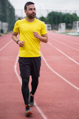 Obraz premium Vibrant young man in a bright t-shirt gracefully runs on an empty track with a red rubber surface and white markings at an outdoor sports ground, healthy lifestyle. 