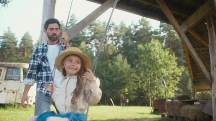 Warm moment of Caucasian dad spending time with his little daughter. Kid with straw hat smiling with joy while having fun on swing. Father looking after beloved child. Vacation in village.