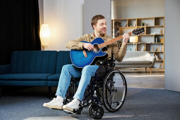 Obraz premium Handsome man with a disability sitting in wheelchair playing on guitar at home