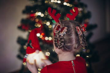 Naklejka premium Blonde girl in a beautiful red dress with a Christmas hairstyle and beautiful hairpins on her head against the background of a Christmas tree