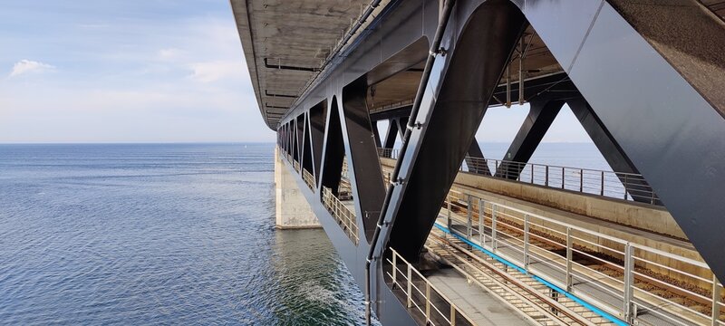 Modern bridge structure with geometric steel beams spanning over a calm sea, showcasing industrial design and engineering precision against a blue ocean backdrop