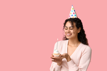 Beautiful young African-American woman in party hat holding birthday cupcake with burning candle on pink background