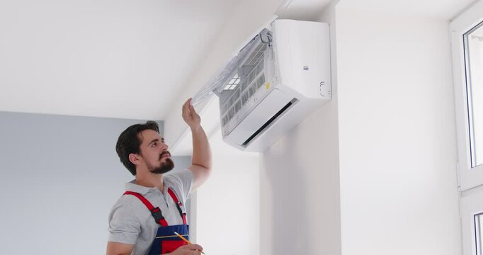 Male engineer repairman in work uniform opening air conditioner panel, inspecting condition of AC system after installing, cleaning or repairing. Technician service and maintenance of cooling systems.
