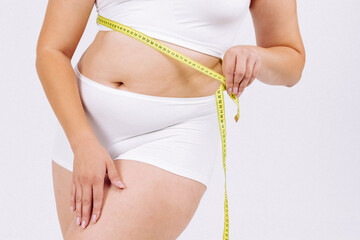 A young plus-size woman measuring her waist against a white background. A concept of weight loss, fitness, and self-confidence, inspiring a healthy lifestyle and goal achievement.