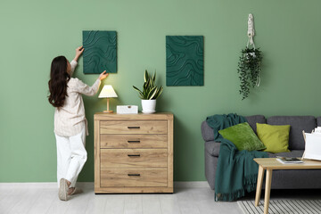 Young pretty woman hanging picture on green wall near chest of drawers in living room