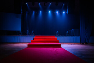 Dramatic scene with a red carpet staircase leading up to a stage, illuminated by blue spotlights....