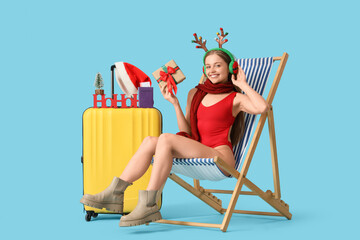 Young woman in swimsuit and reindeer horns with gift box and suitcase sitting in deck chair on blue background. Christmas in July