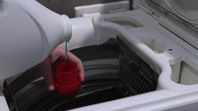 Woman pouring laundry gel into detergent cap in preparation for wash cycle, close-up of washing machine drum