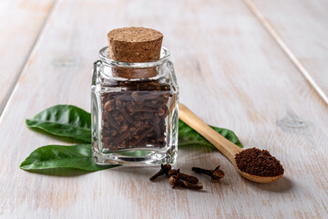 Whole and ground clove buds over white wooden background. Glass jar full of cloves and powdered spice in a wooden spoon close-up. Spice and herbal medicine concept.