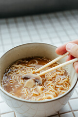 view of spicy instant noodles with pieces of mushrooms, green and red chili peppers and vegetables, spicy ramen noodles on chopsticks in white ceramic bowl. noodle sticks, fast food, hot soup close up
