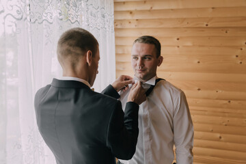 Two men are getting dressed up, one of them is adjusting the tie of the other. The man in the suit is wearing a tie and the other man is adjusting it