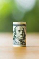 Close-up of a rolled up one hundred dollar bill on a wooden surface with a blurred green background