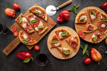 Pizza with pepperoni, basil and pears with glasses of wine, pizza knife, fresh vegetables on cutting boards on a dark background