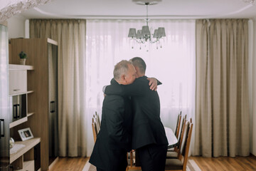 Two men hug each other in a living room with a large dining table in the background. Scene is warm and friendly, as the two men are embracing each other